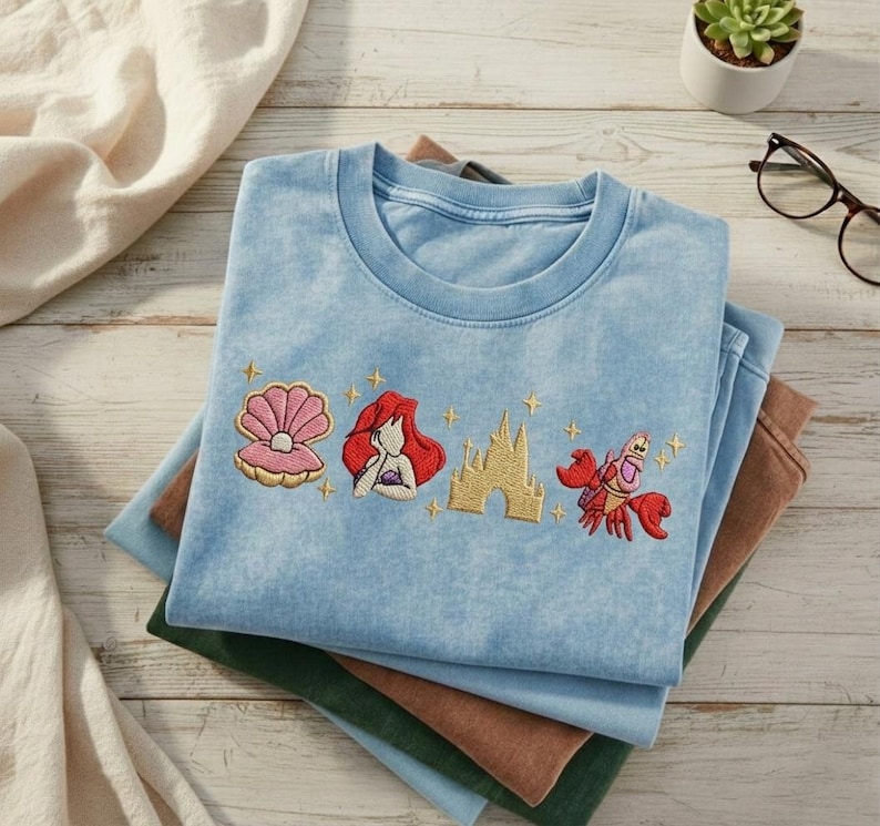 K&ouml;nnte beinhalten: Hellblaues T-Shirt mit gestickten Bildern einer Muschel, einer rothaarigen Frau, einem Schloss und einer roten Krabbe. Das Shirt ist auf anderen gefalteten Shirts in Gr&uuml;n- und Braunt&ouml;nen gefaltet.