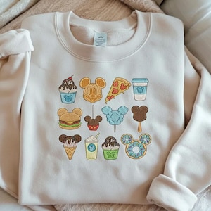 Embroidered Disney snacks sweatshirt with Mickey-shaped food icons including ice cream, donuts, popcorn, coffee, and treats on cream sweatshirt