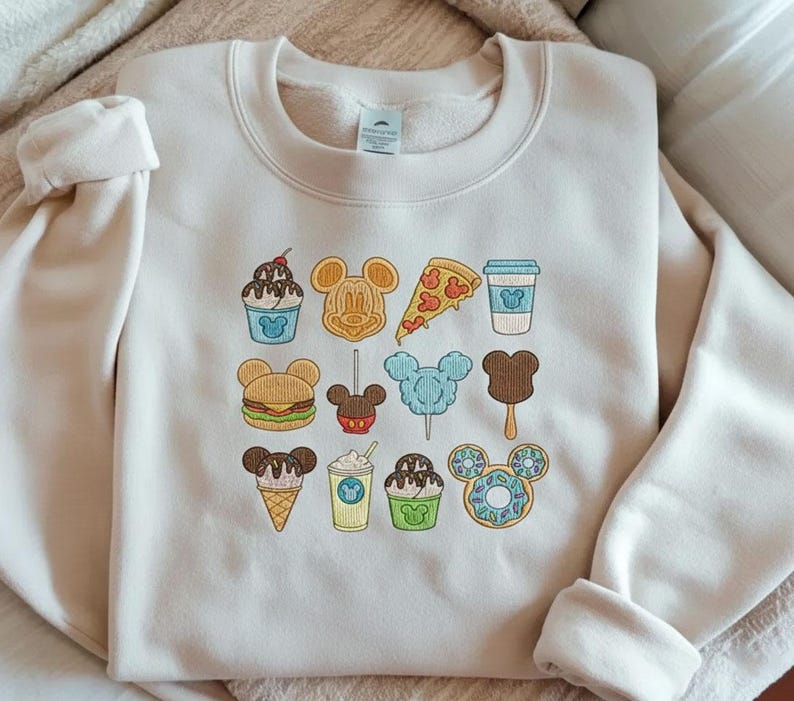 Embroidered Disney snacks sweatshirt with Mickey-shaped food icons including ice cream, donuts, popcorn, coffee, and treats on cream sweatshirt