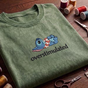 May include: Olive green t-shirt with an embroidered blue cartoon character design and the word "overstimulated" in black. The character is lying down with a floral garland. The shirt is folded on a wooden surface.