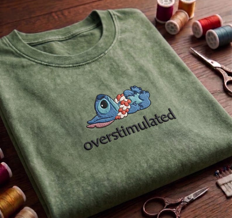 May include: Olive green t-shirt with an embroidered blue cartoon character design and the word "overstimulated" in black. The character is lying down with a floral garland. The shirt is folded on a wooden surface.