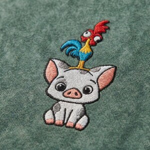 May include: Embroidered design featuring a cartoon pig with a rooster perched on its head. The pig is white with black spots and pink ears and snout. The rooster is red, blue, and yellow. The design is on a green fabric.