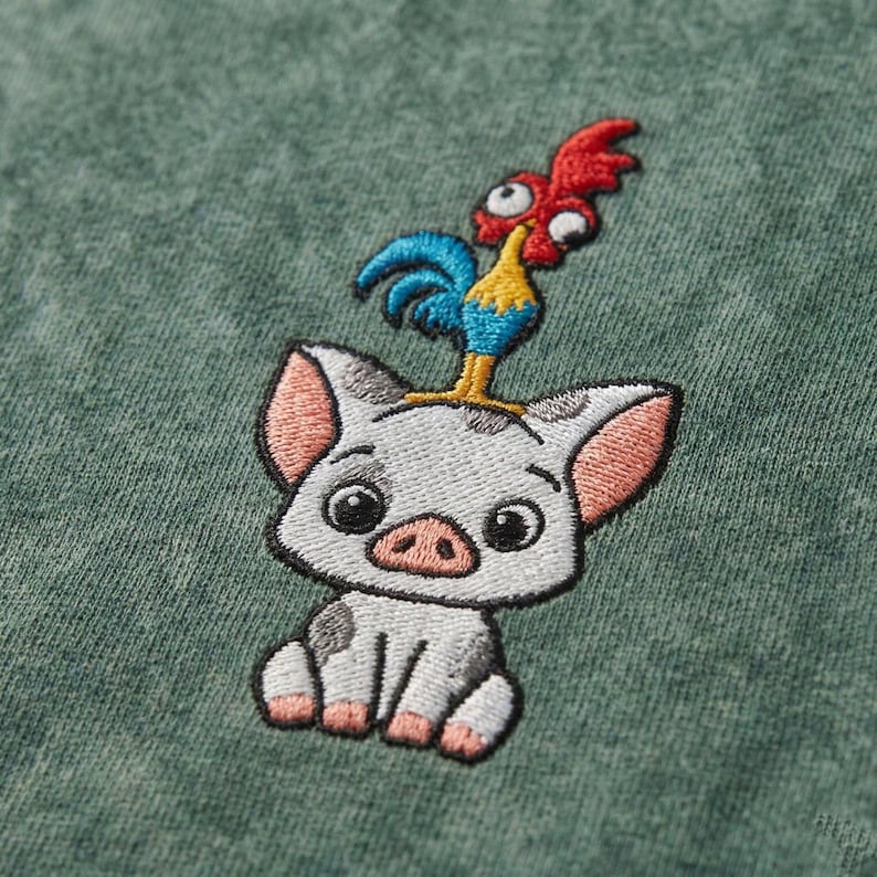 May include: Embroidered design featuring a cartoon pig with a rooster perched on its head. The pig is white with black spots and pink ears and snout. The rooster is red, blue, and yellow. The design is on a green fabric.