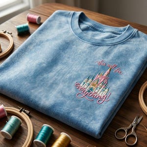 Puede incluir: Una camiseta azul claro con un dise&ntilde;o de castillo bordado y el texto "This will fix everything!" en rosa. Aros de bordado, carretes de hilo y tijeras est&aacute;n dispuestos alrededor de la camiseta sobre una superficie de madera.