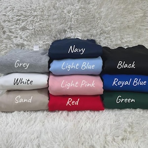 May include: A collection of folded sweatshirts in various colors, including grey, navy, light blue, black, white, light pink, royal blue, sand, red, and green. Each sweatshirt has its color name printed on it in white.