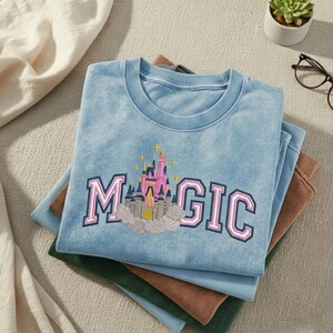 May include: A light blue t-shirt with the word "MAGIC" in pink and blue lettering, featuring an embroidered castle design. The shirt is folded on top of other shirts in shades of blue, brown, and green.
