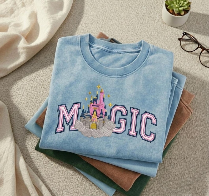 May include: A light blue t-shirt with the word "MAGIC" in pink and blue lettering, featuring an embroidered castle design. The shirt is folded on top of other shirts in shades of blue, brown, and green.