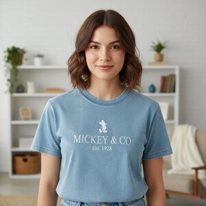 May include: A light blue t-shirt featuring the text "MICKEY & CO Est. 1928" in white, along with a small Mickey Mouse graphic. The shirt is a crew neck style and is being worn by a person.