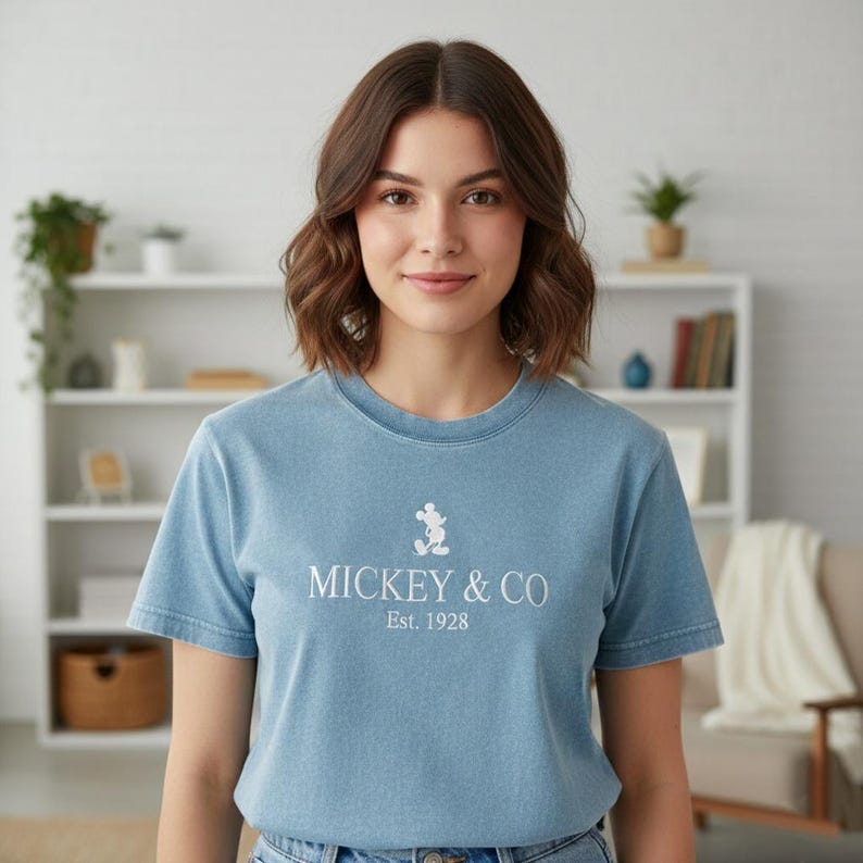 May include: A light blue t-shirt featuring the text "MICKEY & CO Est. 1928" in white, along with a small Mickey Mouse graphic. The shirt is a crew neck style and is being worn by a person.
