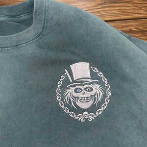 Op de afbeelding: Tealkleurige crewneck sweatshirt met een wit geborduurd ontwerp. Het ontwerp is een griezelige, grijnzende schedel met een hoge hoed, omringd door een decoratieve ketting. De sweatshirt is gemaakt van zacht materiaal.