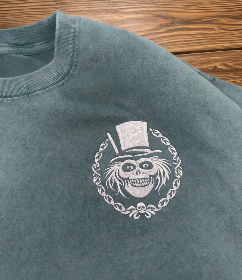 Op de afbeelding: Tealkleurige crewneck sweatshirt met een wit geborduurd ontwerp. Het ontwerp is een griezelige, grijnzende schedel met een hoge hoed, omringd door een decoratieve ketting. De sweatshirt is gemaakt van zacht materiaal.