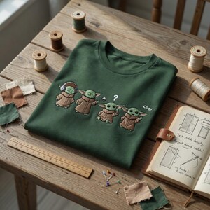 Puede incluir: Camiseta verde oscuro de cuello redondo con un dise&ntilde;o bordado de cuatro personajes de Grogu en varias poses. El dise&ntilde;o incluye la palabra "cool!" y un signo de interrogaci&oacute;n. La camiseta est&aacute; doblada sobre una superficie de madera con suministros de costura.