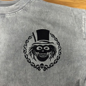 Op de afbeelding: Lichtgrijs t-shirt met een zwart geborduurd ontwerp van een schedel met hoge hoed, omringd door een decoratieve krans. Het ontwerp is gecentreerd op de borst van het shirt.