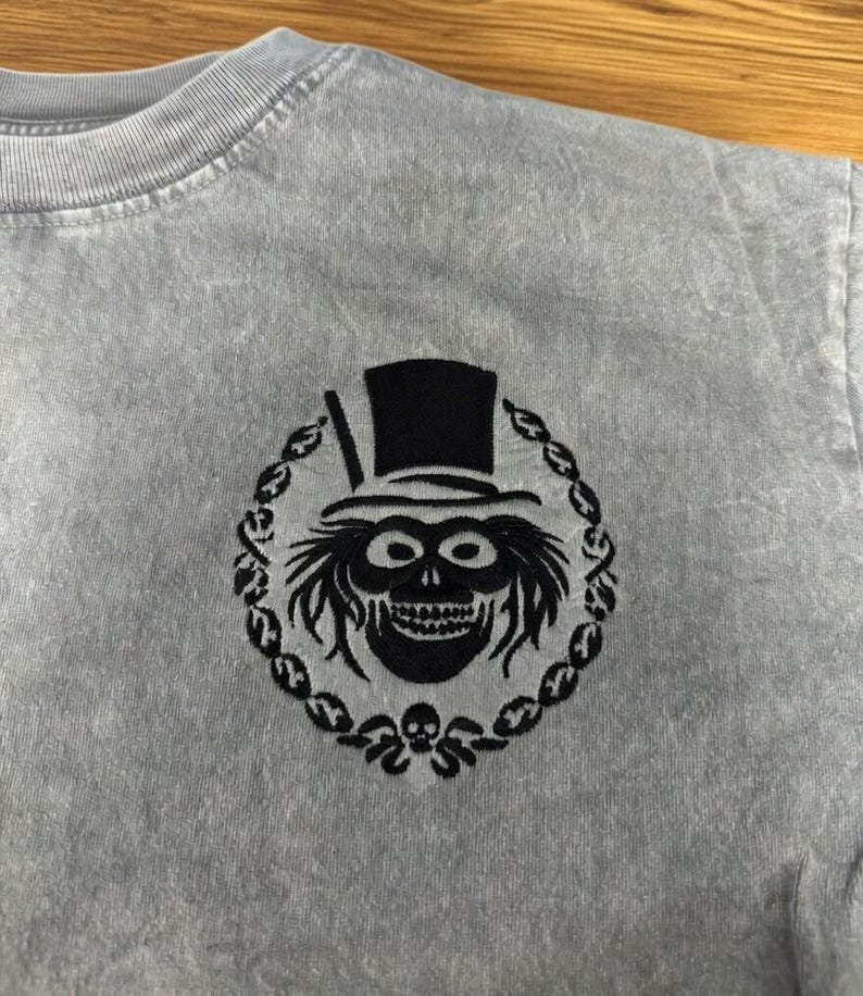 Op de afbeelding: Lichtgrijs t-shirt met een zwart geborduurd ontwerp van een schedel met hoge hoed, omringd door een decoratieve krans. Het ontwerp is gecentreerd op de borst van het shirt.