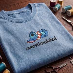 May include: Light blue t-shirt with an embroidered cartoon character and the word "overstimulated" in black. The character is blue with a large eye and a lei. Sewing supplies, including thread spools and scissors, are in the background.