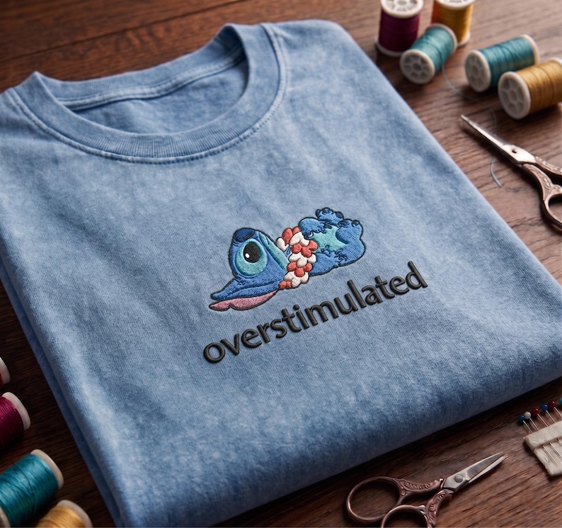 May include: Light blue t-shirt with an embroidered cartoon character and the word "overstimulated" in black. The character is blue with a large eye and a lei. Sewing supplies, including thread spools and scissors, are in the background.