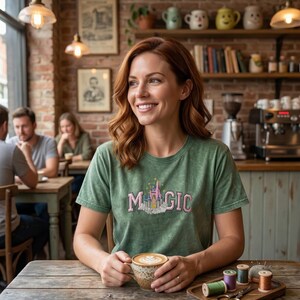 May include: A green t-shirt with the word "MAGIC" in pink and a castle graphic. The person is holding a cup of coffee with latte art. The scene is set in a cafe with a wooden table and brick wall.