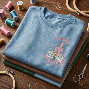 Puede incluir: Una pila de tres camisetas en azul, marr&oacute;n y verde. La camiseta azul superior presenta un dise&ntilde;o de castillo rosa bordado con el texto "this will fix everything!". Suministros de bordado, incluyendo carretes de hilo y tijeras, est&aacute;n dispuestos alrededor de las camisas.