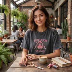May include: A woman wearing a dark gray t-shirt with the text "Happiest Mom On Earth" and a pink castle graphic. She is sitting at a wooden table with a coffee cup, scissors, and a book. The setting is a cafe.