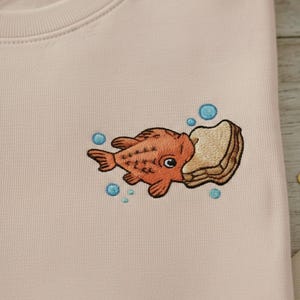 May include: A light pink sweatshirt featuring an embroidered design of an orange fish biting into a piece of bread. Blue bubbles surround the fish and bread. The sweatshirt is folded.