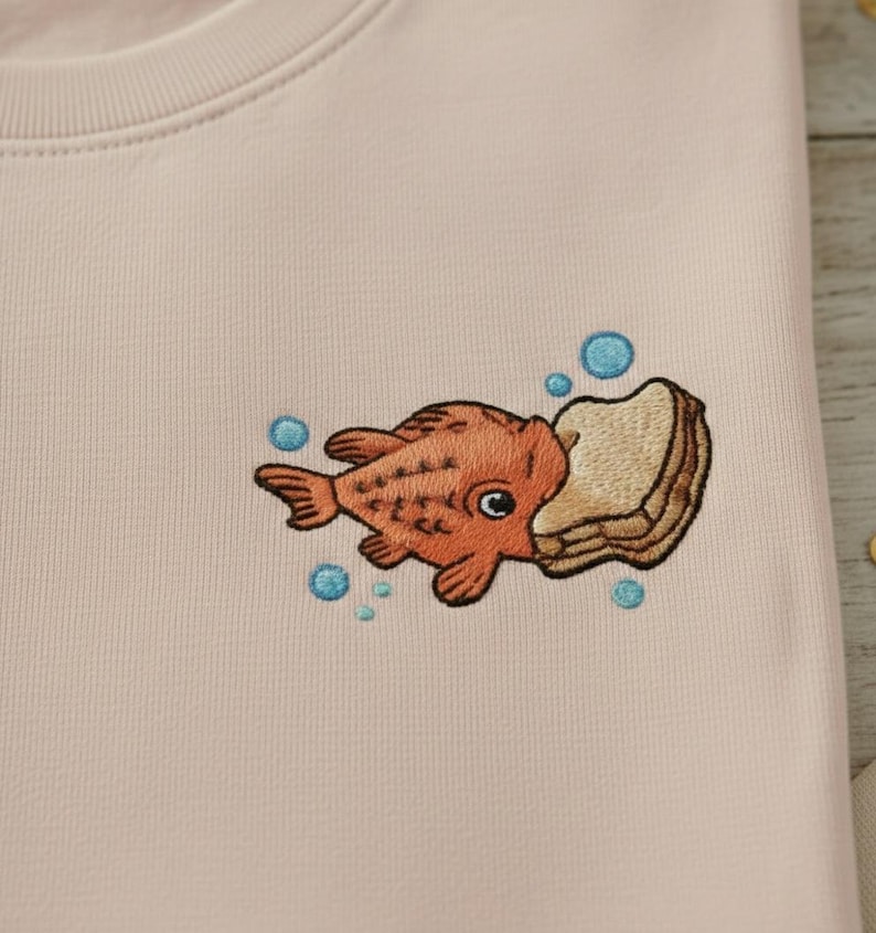 May include: A light pink sweatshirt featuring an embroidered design of an orange fish biting into a piece of bread. Blue bubbles surround the fish and bread. The sweatshirt is folded.