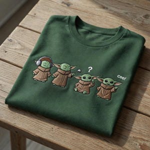 Puede incluir: Camiseta verde oscuro con un dise&ntilde;o bordado de un personaje en varias poses. El personaje lleva auriculares, tiene un signo de interrogaci&oacute;n sobre la cabeza y la palabra "cool!" est&aacute; al lado.