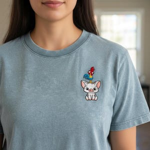 May include: A light blue t-shirt with an embroidered design of a pig with a rooster on its head. The pig is white with gray spots and the rooster is red and blue. The shirt has a crew neck and short sleeves.