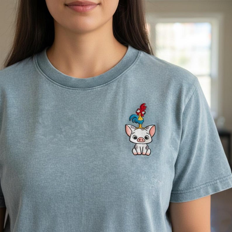 May include: A light blue t-shirt with an embroidered design of a pig with a rooster on its head. The pig is white with gray spots and the rooster is red and blue. The shirt has a crew neck and short sleeves.