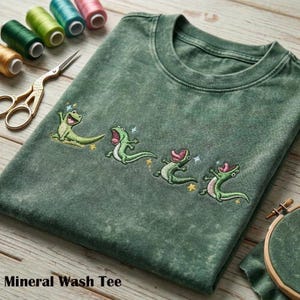 Puede incluir: Camiseta verde lavado mineral con bordados de lagartos de dibujos animados. Los lagartos están en varias poses con la boca abierta y estrellas brillantes. El texto "Mineral Wash Tee" está en la parte inferior de la imagen.
