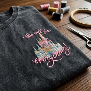 Puede incluir: Camiseta gris oscuro con un dise&ntilde;o de castillo bordado y la frase "this will fix everything!" en escritura rosa. El castillo presenta detalles en rosa, azul y dorado. Suministros de bordado, incluyendo hilo y tijeras, en el fondo.