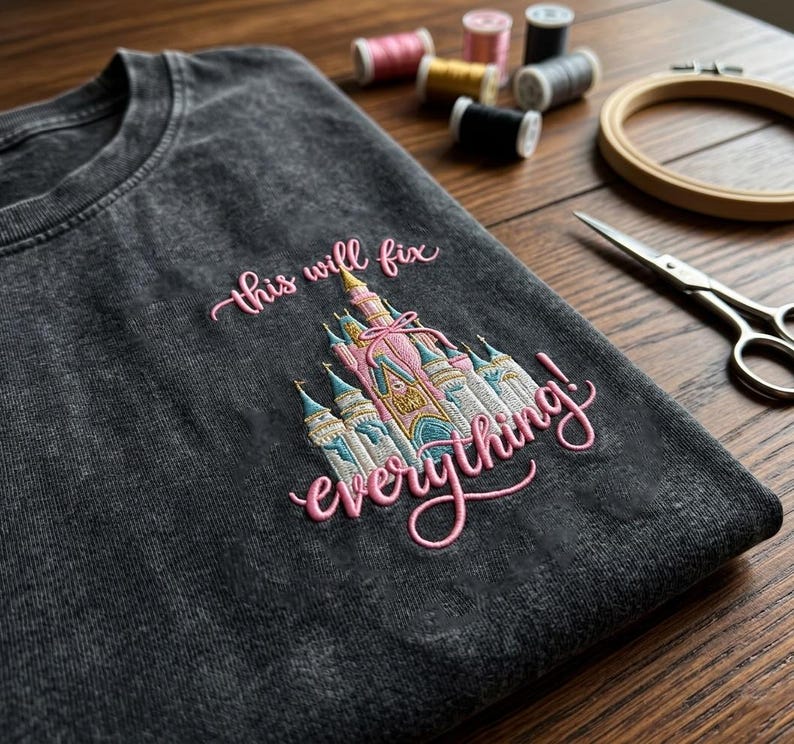Puede incluir: Camiseta gris oscuro con un dise&ntilde;o de castillo bordado y la frase "this will fix everything!" en escritura rosa. El castillo presenta detalles en rosa, azul y dorado. Suministros de bordado, incluyendo hilo y tijeras, en el fondo.