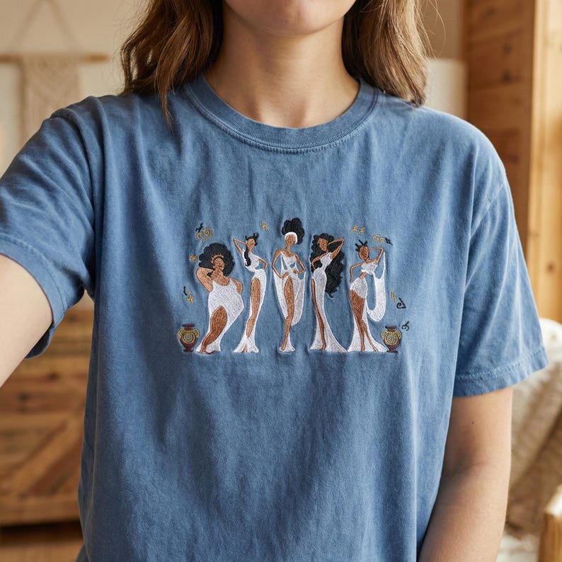 Peut inclure: Un t-shirt bleu avec une broderie repr&eacute;sentant cinq figures stylis&eacute;es en robes blanches. Les figures ont diff&eacute;rentes coiffures et des d&eacute;tails dor&eacute;s. Le t-shirt est fait d'une mati&egrave;re douce et confortable.