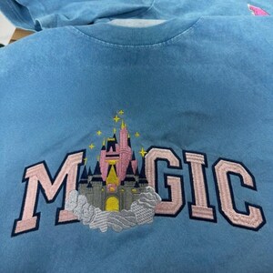 May include: Light blue t-shirt with the word "MAGIC" embroidered in pink, featuring a castle design. The castle has pink and gray towers with a yellow entrance, and is surrounded by clouds and stars.
