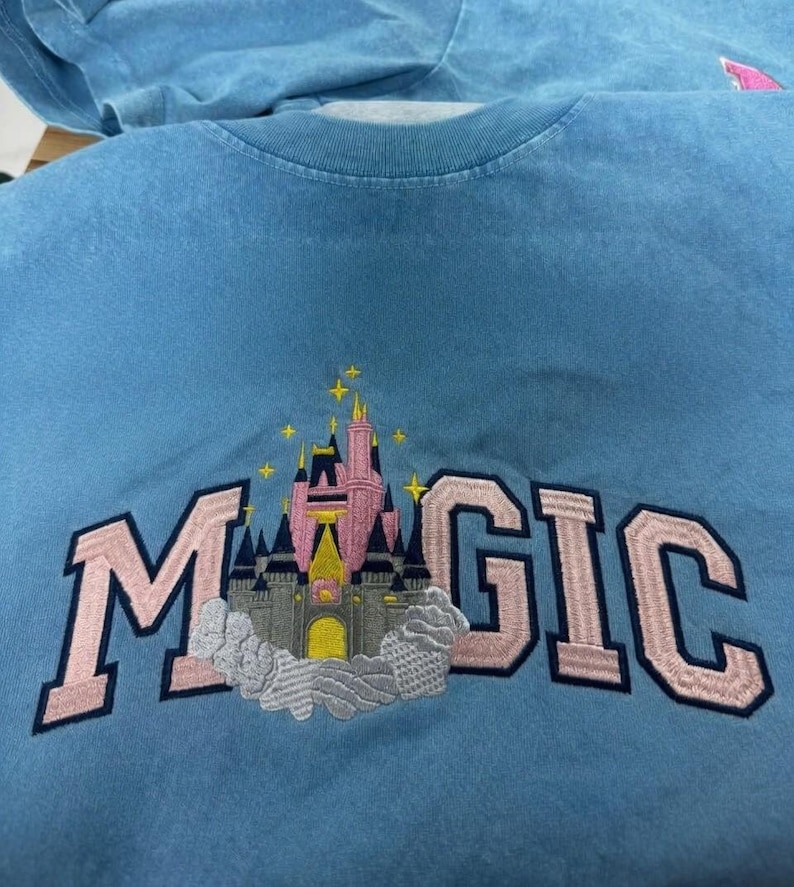 May include: Light blue t-shirt with the word "MAGIC" embroidered in pink, featuring a castle design. The castle has pink and gray towers with a yellow entrance, and is surrounded by clouds and stars.