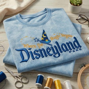 Op de afbeelding: Lichtblauwe t-shirts met geborduurd "Disneyland Park" ontwerp. Het ontwerp bevat een tovenaars hoed, ster en de tekst "Where the Magic Began". De shirts zijn op een houten oppervlak gevouwen met naai benodigdheden.
