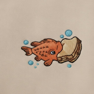 May include: Embroidered design of an orange goldfish eating a piece of bread, with blue bubbles around it. The design is on a beige fabric. The goldfish has a large eye and detailed fins.