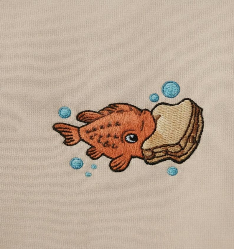 May include: Embroidered design of an orange goldfish eating a piece of bread, with blue bubbles around it. The design is on a beige fabric. The goldfish has a large eye and detailed fins.