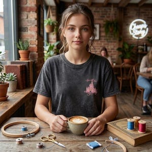 Puede incluir: Una mujer con una camiseta gris oscuro con un gr&aacute;fico de castillo rosa y el texto "this will be everything". Sostiene una taza de caf&eacute; con arte latte. Suministros de bordado y un libro est&aacute;n sobre la mesa de madera.