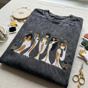 Peut inclure: T-shirt gris fonc&eacute; avec une broderie repr&eacute;sentant cinq figures stylis&eacute;es en robes blanches et dor&eacute;es. Les figures ont des cheveux noirs et sont dans diff&eacute;rentes poses. Des fournitures de broderie sont visibles.