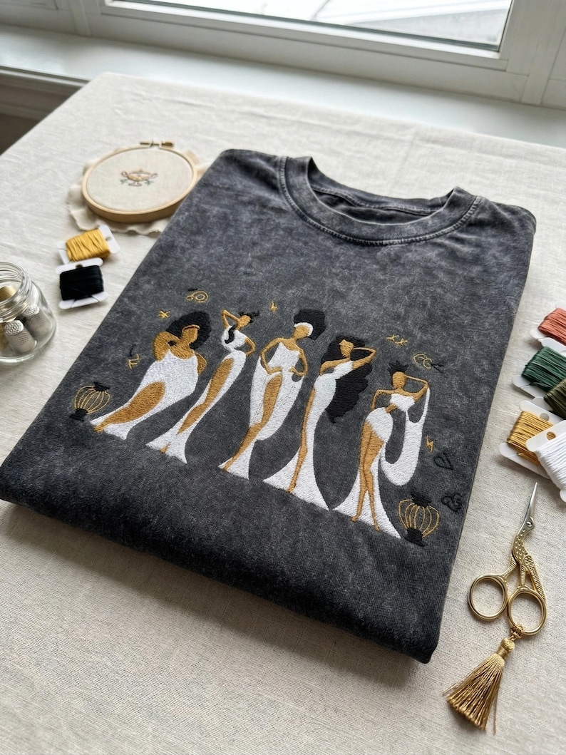 Peut inclure: T-shirt gris fonc&eacute; avec une broderie repr&eacute;sentant cinq figures stylis&eacute;es en robes blanches et dor&eacute;es. Les figures ont des cheveux noirs et sont dans diff&eacute;rentes poses. Des fournitures de broderie sont visibles.