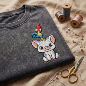 May include: Dark gray crew neck t-shirt with an embroidered design featuring a cartoon pig with a rooster on its head. The pig is white with pink ears and blue spots. Sewing supplies, including thread spools and pins, are in the background.