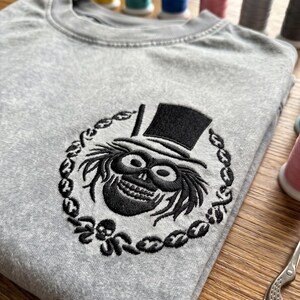 Op de afbeelding: Een grijze crewneck sweatshirt met een zwart geborduurd ontwerp van een schedel met hoge hoed, omringd door een kettingachtige rand. Het ontwerp is gecentreerd op de borst. Spoelen garen zijn zichtbaar op de achtergrond.