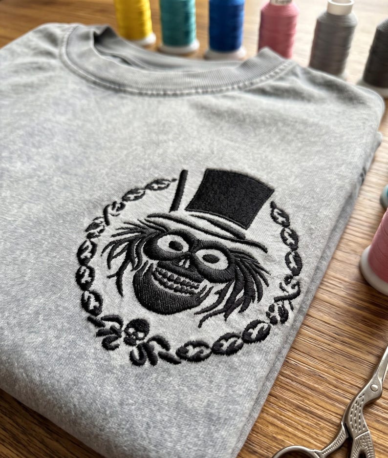 Op de afbeelding: Een grijze crewneck sweatshirt met een zwart geborduurd ontwerp van een schedel met hoge hoed, omringd door een kettingachtige rand. Het ontwerp is gecentreerd op de borst. Spoelen garen zijn zichtbaar op de achtergrond.