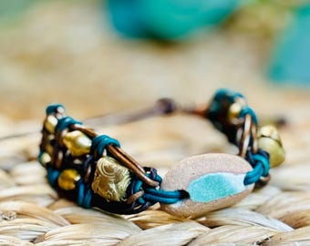 May include: A handcrafted bracelet featuring a central stone with turquoise accents, complemented by gold-toned beads and dark blue leather accents. The bracelet is made with brown leather and has a natural, earthy aesthetic.
