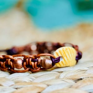May include: Close-up of a handmade bracelet featuring brown leather, copper beads, and a yellow, ridged ceramic bead. The bracelet is intricately woven with purple accents, set against a natural woven background.