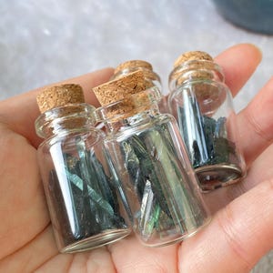 Op de afbeelding: Kleine glazen flesjes met kurken stoppen, elk gevuld met donkerblauwe-groene kristalfragmenten. De flesjes worden in een hand gehouden, waarbij de kleur en textuur van de kristallen worden benadrukt. Het heldere glas maakt de inhoud zichtbaar.