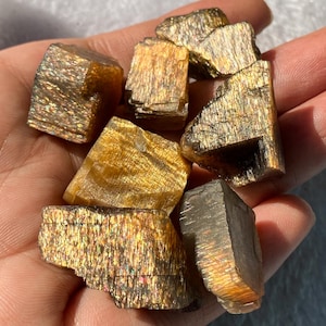 Puede incluir: Una colección de cristales de piedra solar en bruto, que muestran una iridiscencia dorada-naranja brillante. Las piedras en bruto tienen un aspecto natural y texturizado, con estructuras cristalinas visibles. Los cristales se sostienen en una mano, mostrando su color y brillo únicos.