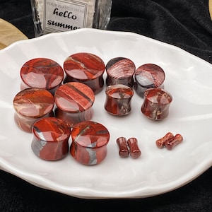 May include: A collection of red and brown stone ear plugs and gauges in various sizes, displayed on a white, scalloped plate. The plugs have a marbled appearance, with a mix of red and brown tones. The image also includes a small succulent in a glass container with the text "hello summer".