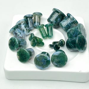 Könnte beinhalten: Eine Sammlung grüner und blauer Stein-Ohrstecker in verschiedenen Größen und Formen. Die Ohrstecker sind auf einem weißen, quadratischen Keramiktablett angeordnet. Der Stein hat ein marmoriertes Aussehen mit grünen und blauen Wirbeln.