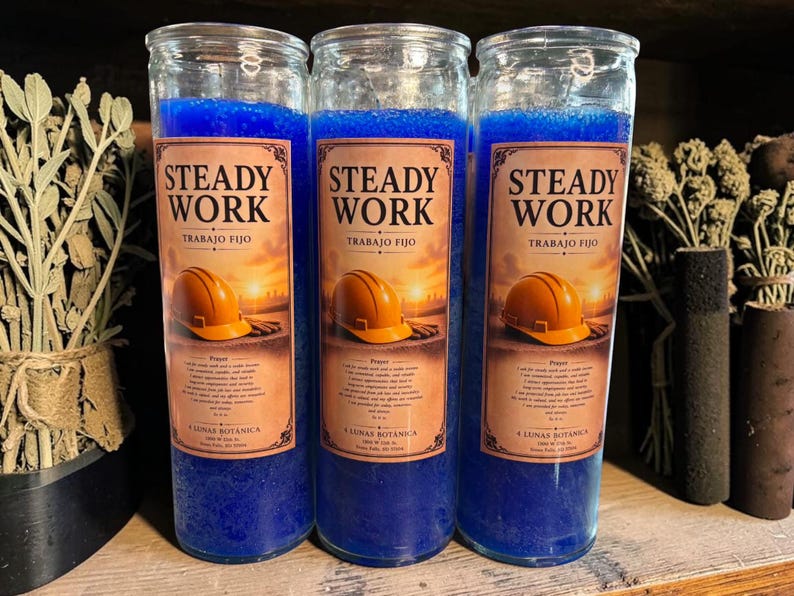 Puede incluir: Tres velas cil&iacute;ndricas altas de vidrio rellenas de cera azul vibrante. Cada vela presenta una etiqueta con el texto STEADY WORK, TRABAJO FIJO, una ilustraci&oacute;n de un casco de obra naranja sobre un atardecer y un texto de oraci&oacute;n.